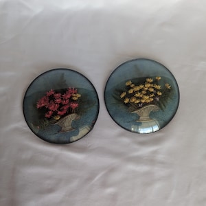 May include: Two circular wall plaques featuring convex glass covers. One plaque displays pink flowers in a tan basket, while the other shows yellow flowers in a matching basket. Both have a blue background and a thin dark metal rim around the edge.