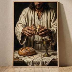 May include: A framed wall art print depicting a figure with long hair and a beard wearing a cream-colored robe. The figure is breaking a loaf of bread with both hands over a white cloth, with a whole loaf and a gold chalice resting on the table.