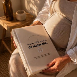 May include: A beige binder with a clear front cover featuring the text "mon dossier de maternit&eacute;." and a small illustration of two baby feet. The binder includes additional French text detailing its use for appointments and exams.