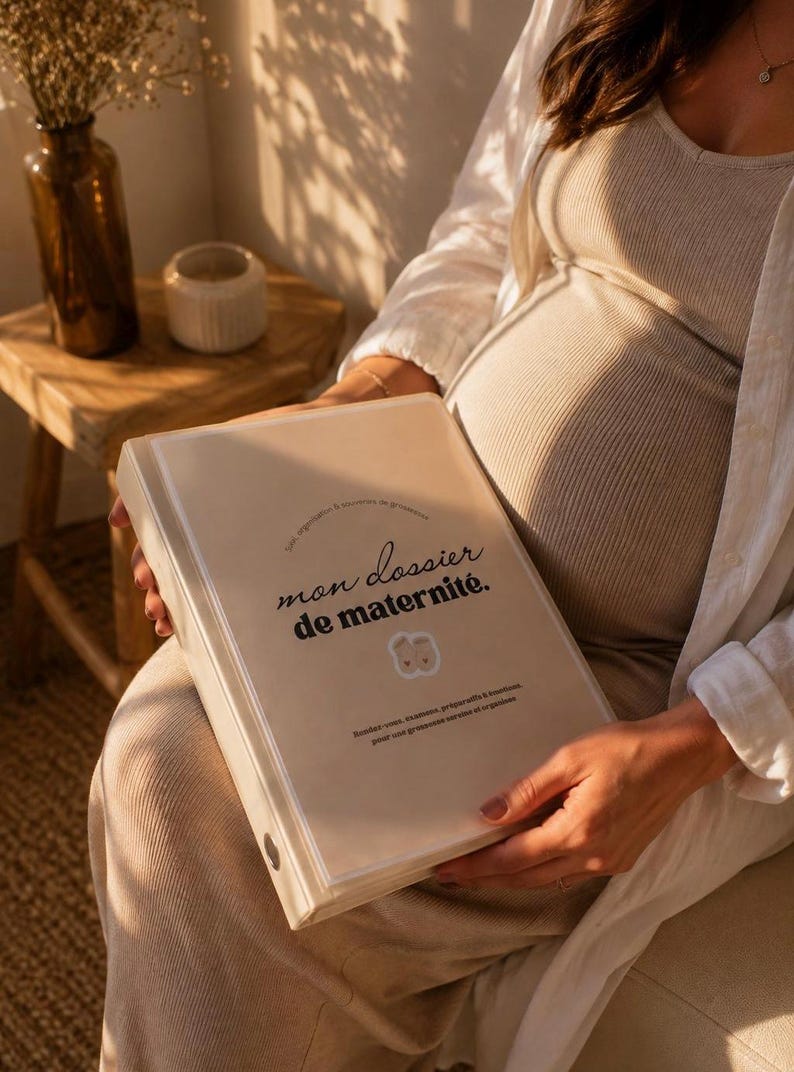 May include: A beige binder with a clear front cover featuring the text "mon dossier de maternit&eacute;." and a small illustration of two baby feet. The binder includes additional French text detailing its use for appointments and exams.