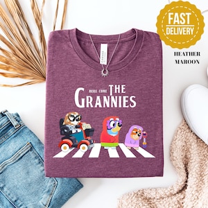 May include: A heather maroon short-sleeved t-shirt featuring a graphic of three cartoon dogs dressed as elderly characters on a crosswalk. The text above the characters reads Here Come The Grannies. The shirt has a crew neckline and a white brand label.