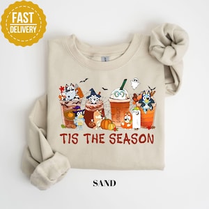 May include: A sand-colored crewneck sweatshirt featuring a graphic of cartoon dogs in Halloween costumes inside coffee cups, with pumpkins, bats, and spiderwebs. The text "TIS THE SEASON" is printed in brown below the graphic. The fabric is a light beige.
