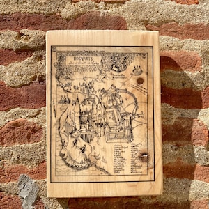 May include: A wooden plaque featuring a detailed black and white map of Hogwarts. The map is framed by a light-colored wood and mounted on a brick wall. The text "HOGWARTS" is visible at the top of the map.