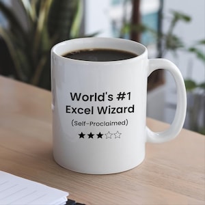 May include: A white ceramic mug featuring the black printed text World's #1 Excel Wizard (Self-Proclaimed) above three solid black stars and two outlined stars. The mug is filled with dark coffee and has a standard handle on the right side.