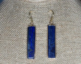 May include: A pair of drop earrings featuring rectangular lapis lazuli stones in a deep blue hue with natural dark inclusions. Each stone is encased in a thin gold-toned metal frame and suspended from a simple gold-toned curved ear wire hook.