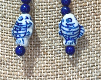 May include: A pair of dangle earrings featuring silver-toned hooks, three small round deep blue beads, and a central white ceramic fish-shaped bead with blue hand-painted striped patterns, finished with a single small round deep blue bead at the base.