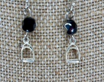 May include: A pair of silver-toned drop earrings featuring a small black faceted bead suspended above a miniature metal stirrup charm. The earrings are attached to simple silver-toned ear wires for hanging, showcasing a equestrian-themed design.