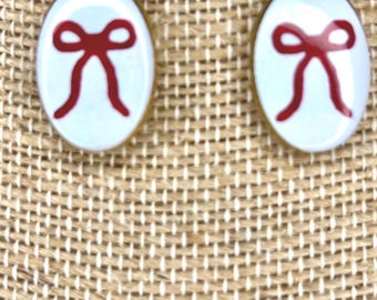 May include: A pair of oval-shaped drop earrings featuring a smooth, light blue enamel surface with a centered, hand-painted red bow graphic. The earrings are suspended from gold-toned metal hooks, showcasing a clean, minimalist design aesthetic.