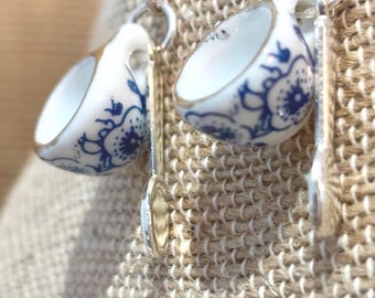 May include: A pair of dangle earrings featuring miniature white ceramic teacups with blue floral patterns and gold-toned rims. Each cup is paired with a small silver-toned spoon charm, all suspended from silver-toned metal earring hooks.