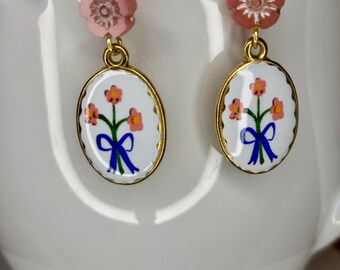 May include: A pair of dangle earrings featuring a small pink flower bead at the top, connected to an oval-shaped charm with a white background. The charm displays a painted bouquet of three orange flowers tied with a blue ribbon in a gold frame.