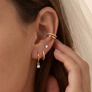 May include: A set of gold-toned earrings consisting of a double-band ear cuff with small clear stones, a single stud earring, and a small hoop earring featuring a dangling clear stone charm. The jewelry is worn on the earlobe and cartilage.