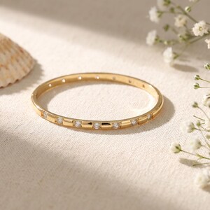 May include: A thin gold bangle bracelet featuring a series of small, round, clear stones set at regular intervals along the outer surface. The metal has a polished finish, and the design includes a hinged closure mechanism visible on the side.