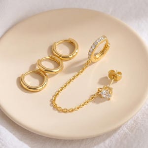 May include: A set of gold-toned jewellery consisting of three plain hoop earrings and one double-piercing earring. The double-piercing earring features a hoop with embedded clear stones connected by a gold chain to a single stud with a clear stone.