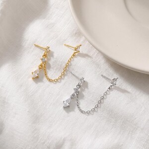 May include: Two double-post chain earrings, one in gold tone and one in silver tone. Each earring features two posts connected by a delicate chain, with a small star-shaped accent and a dangling marquise-cut clear stone charm attached to one post.