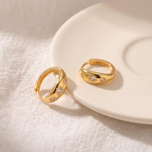 May include: A pair of gold hoop earrings featuring a polished finish and engraved sunburst motifs. Each sunburst design is centered with a small, clear, round-cut gemstone. The earrings have a hinged closure mechanism for secure wear.