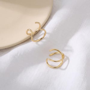 May include: Two gold-toned ear cuffs featuring a smooth, polished finish. Each piece is designed with a double-loop, open-ended structure that curves into a rounded shape, creating a continuous, fluid line that rests securely on the ear cartilage.