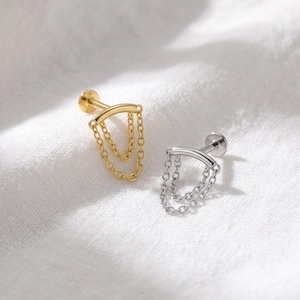 Puede incluir: Dos piercings labret, uno dorado y otro plateado, cada uno con una barra curva y dos delicadas cadenas colgantes unidas a los extremos. Se aprecian los postes de base plana y las cadenas crean un efecto drapeado y superpuesto.