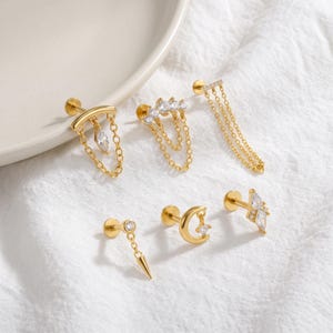 May include: A set of six gold-tone stud earrings featuring various designs including dangling chains, a crescent moon, a spike, and clear marquise-cut stones. Each piece is crafted with a flat back post and incorporates small clear crystals.