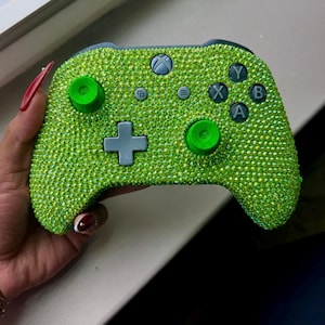 May include: A lime green Xbox controller covered in sparkling rhinestones. The controller features the standard button layout with a directional pad, face buttons, and shoulder buttons. The buttons and joysticks are also green.