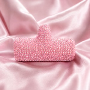 May include: A pink, bedazzled phone grip, covered in small, iridescent crystals. The grip is a light pink color and is set against a backdrop of pink satin fabric.