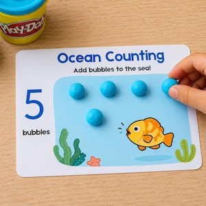 Op de afbeelding: Een witte rechthoekige educatieve kaart met de titel Ocean Counting, de tekst Add bubbles to the sea en 5 bubbles. De kaart toont een gele vis, groen zeewier en een kleine oranje zeester. Er liggen vijf blauwe bollen van boetseerklei op de kaart.
