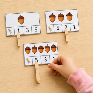 Peut inclure: Trois cartes éducatives rectangulaires illustrées de glands marron. Chaque carte présente une rangée de chiffres 1, 3 et 5, avec une pince à linge en bois fixée pour indiquer une valeur spécifique. Les cartes sont blanches avec du texte noir.