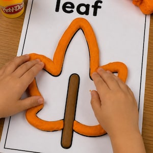May include: A white activity sheet with the word leaf printed in black, featuring an orange playdough outline of a leaf shape and a brown playdough stem. An orange playdough container sits in the corner, and a lump of orange playdough is nearby.