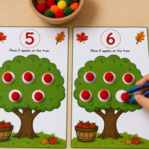 May include: Two educational activity cards featuring illustrations of trees with numbers 5 and 6 at the top. Each card has instructions to place red pom-pom apples on the tree. A wooden bowl contains colorful pom-poms, and blue tweezers are used.