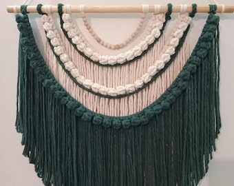 May include: A wall hanging featuring dark green and cream-colored cotton cord arranged in a layered, arched pattern. The design includes rows of knotted texture, a strand of light wooden beads, and a long fringe, all suspended from a wooden dowel.