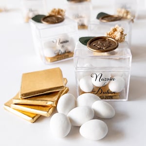May include: Clear square plastic favour boxes containing white sweets, each topped with a gold wax seal, a green leaf, and dried flowers. The boxes are printed with the text Nazrin & Duhan 05.2026. Beside them are gold-wrapped square chocolates.