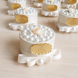Op de afbeelding: Een ronde gebroken witte geschenkdoos met een cluster van witte kunstparels en een centrale crèmekleurige stoffen roos. De zijkant heeft een goudkleurig geschulpt label met de tekst Ayçin & Murat Bir Ömür Boyu 27.08.2019 en een witte strik.