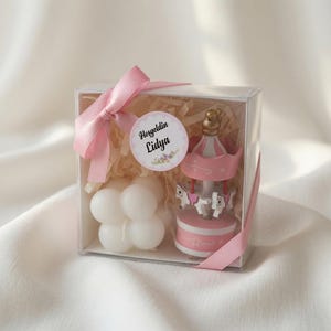 May include: A clear gift box containing a white bubble candle and a pink and white carousel ornament. The box is decorated with a pink satin ribbon and a circular tag featuring the text Horgeldin Lidya. The interior is filled with paper filler.