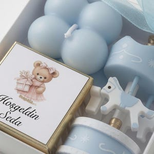 May include: A white gift box containing a light blue bubble candle, a small carousel horse ornament, and a square card featuring a brown teddy bear holding a gift with the text Hosgeldin Seda printed in black script below the illustration.