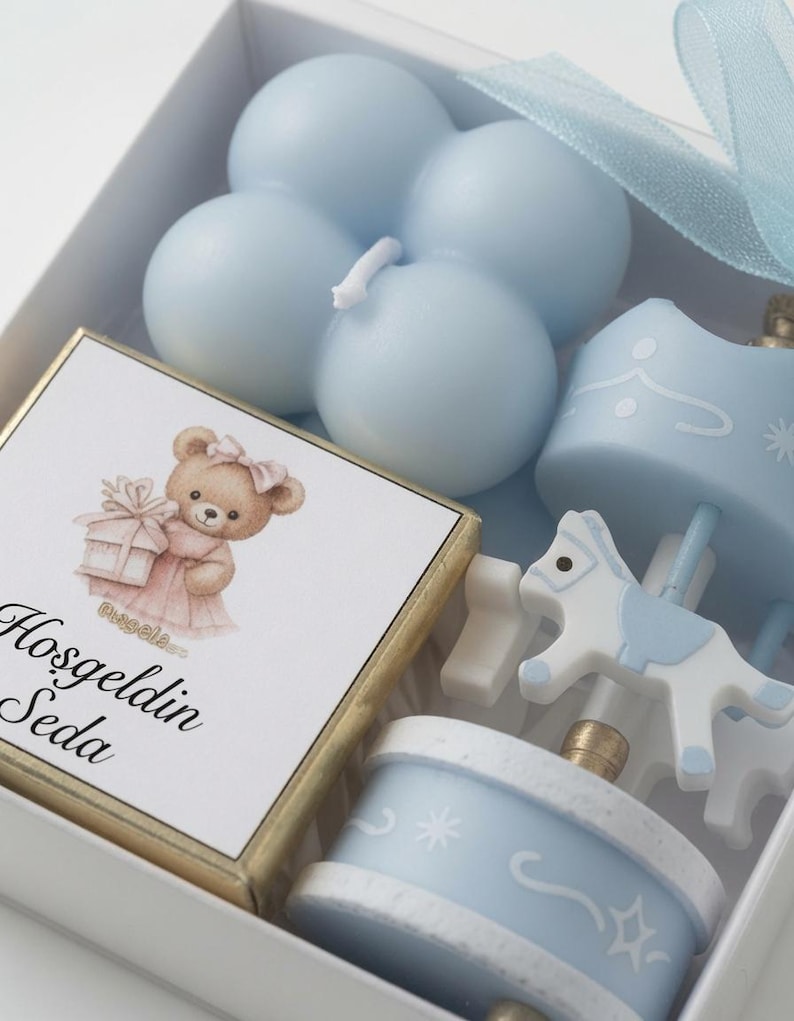May include: A white gift box containing a light blue bubble candle, a small carousel horse ornament, and a square card featuring a brown teddy bear holding a gift with the text Hosgeldin Seda printed in black script below the illustration.