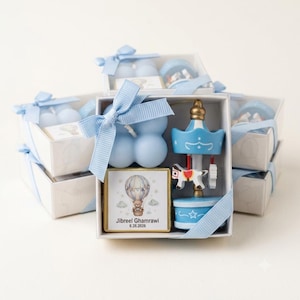 May include: A white gift box containing a light blue bubble candle, a small blue carousel ornament, and a square card featuring a hot air balloon illustration with a teddy bear and the text Jibreel Ghamrawi 6.26.2026, tied with a light blue ribbon.