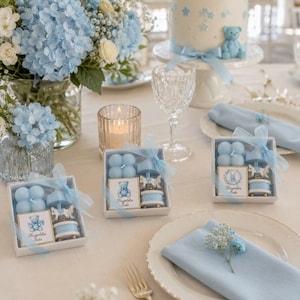 May include: Three square gift boxes containing a light blue bubble candle, a small carousel horse ornament, and a card with a teddy bear illustration and the text Hosgeldin Sola, arranged on a table with blue flowers and napkins.