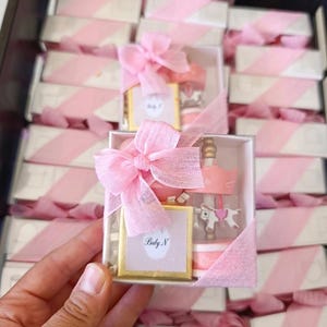 May include: A square white gift box containing a small gold-framed square item with the text Baby N and a miniature pink and white carousel figurine. The box is wrapped with a sheer pink ribbon tied in a bow on the top left corner.