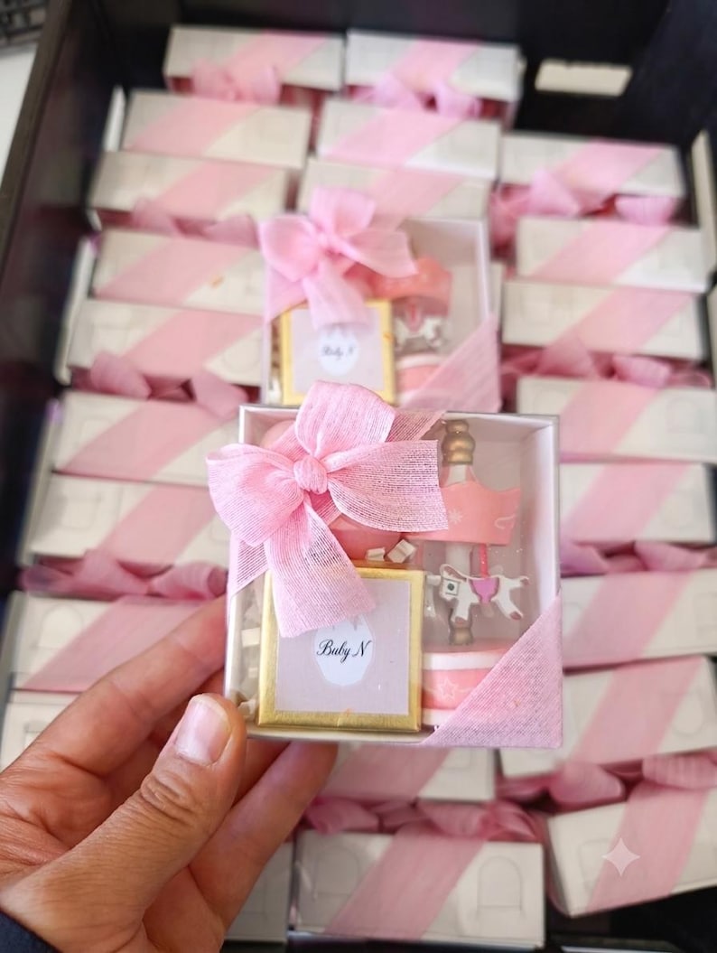 May include: A square white gift box containing a small gold-framed square item with the text Baby N and a miniature pink and white carousel figurine. The box is wrapped with a sheer pink ribbon tied in a bow on the top left corner.