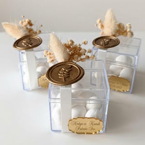 Könnte beinhalten: Drei quadratische, transparente Gastgeschenkboxen gefüllt mit weißen Bonbons. Jede Box ist mit einem weißen Band umwickelt, verziert mit einem goldenen Wachssiegel mit Blattmotiv, beigen Trockenblumen und einem goldenen Namensschild Neelya vs Kamdi.