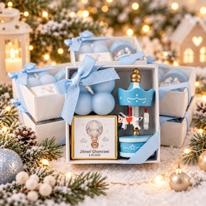 May include: A white gift box tied with a light blue ribbon containing a light blue bubble candle, a small blue and white carousel ornament, and a square chocolate bar with a printed hot air balloon, a teddy bear, and the text Jibreel Ghamrawi 6.26.2026.