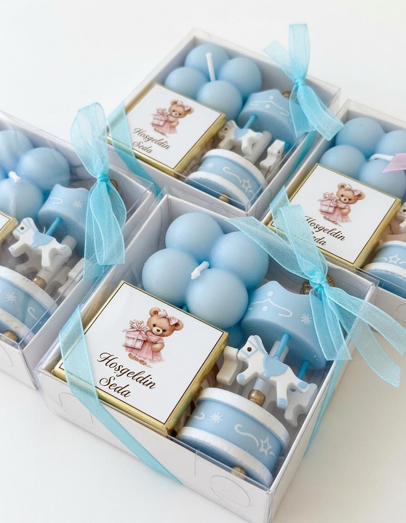 May include: A set of gift boxes containing light blue bubble candles, a carousel horse ornament, and a square chocolate bar with a label featuring a teddy bear illustration and the text Hosgeldin Seda, tied with a light blue sheer ribbon.