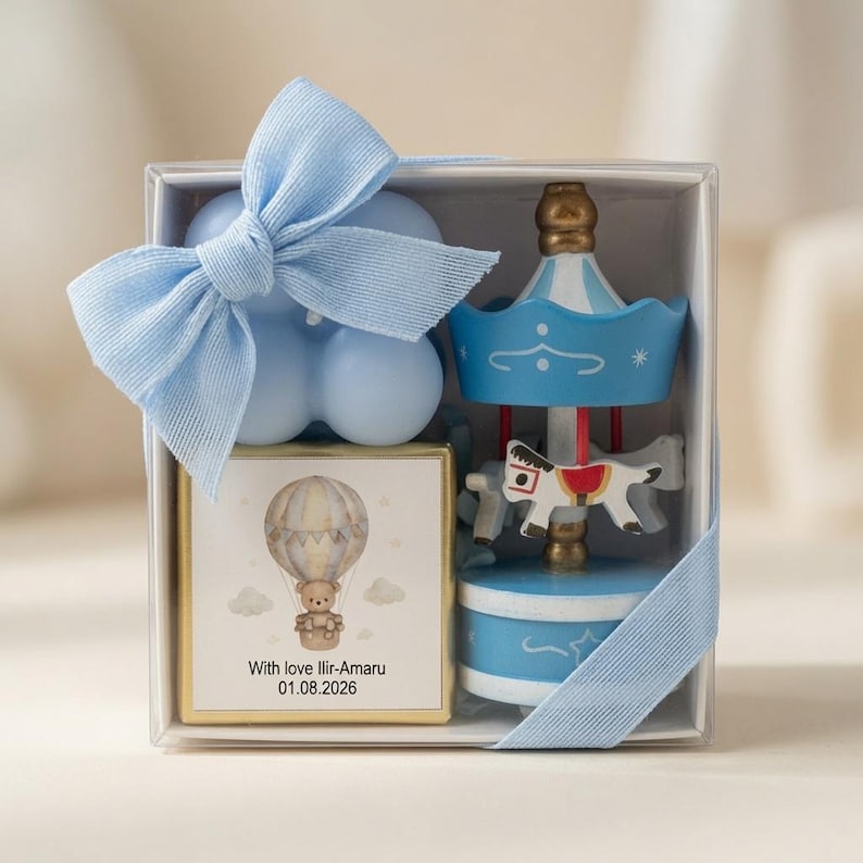 May include: A clear gift box containing a light blue bubble candle with a blue ribbon, a square chocolate bar with a hot air balloon and bear illustration labelled With love Ilir-Amaru 01.08.2026, and a small blue and white carousel music box.