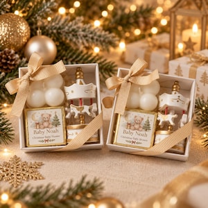 May include: Two white gift boxes each containing a cream-coloured bubble candle, a miniature white carousel with gold accents, and a square card featuring a teddy bear illustration with the text Baby Noah Christmas Baby Shower, tied with a gold ribbon.