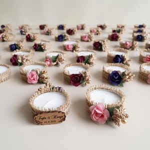 Pode incluir: Uma coleção de velas redondas brancas envolvidas em fita de juta desfiada. Cada vela é decorada com uma pequena rosa artificial em tons de rosa, vermelho ou roxo e bagas douradas. Uma etiqueta de madeira diz Tugba & Hüseyin 02.11.2020.