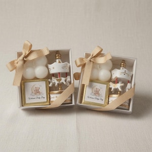 May include: Two square gift boxes each containing a white bubble candle, a small carousel ornament with a horse, and a square card featuring a teddy bear on a crescent moon with the text Welcome Baby Boy, all tied with a beige ribbon.