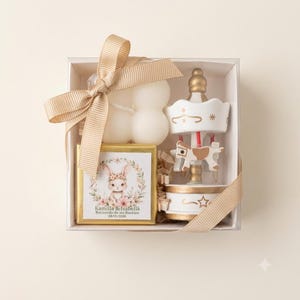 May include: A gift box containing a white bubble candle, a gold-framed square card with a bunny illustration and the text Kamilla & Isabella Recuerdo de mi Bautizo 08/01/2026, and a white and gold carousel ornament, all tied with a beige ribbon.