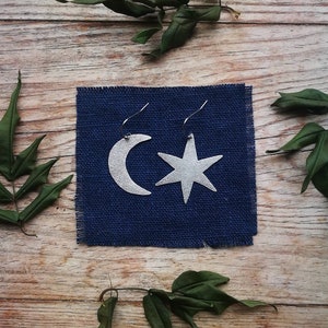 Puede incluir: Un par de pendientes plateados, uno con forma de media luna y el otro de estrella de cinco puntas, cuelgan de un cuadrado de tela azul. Los pendientes están sobre un fondo de madera, con hojas verdes esparcidas.