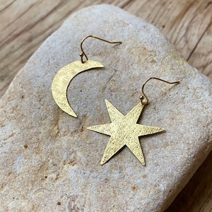 Op de afbeelding: Een paar goudkleurige oorbellen, een in de vorm van een halve maan en de andere een zespuntige ster. Beide oorbellen hebben een getextureerd oppervlak en worden gepresenteerd op een lichtgekleurde steen. De oorbellen hebben een haaksluiting.