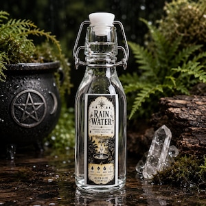May include: A clear glass swing-top bottle filled with liquid, featuring a rectangular label with the text RAIN WATER, NATURALLY COLLECTED DURING RAINFALL, and ALIEN ALOE LLC. The label includes black and white illustrations of a forest and cauldron.