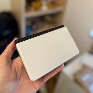 Puede incluir: Una consola de juegos portátil con exterior blanco y parte superior negra. El dispositivo es rectangular con esquinas redondeadas y se sostiene en la mano de una persona. La pantalla es blanca y la parte superior es negra.